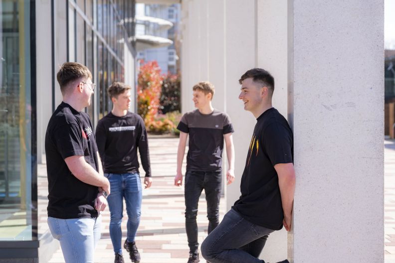 Two young men standing facing each other and chatting outside the university as two other young men walking towards them also chatting